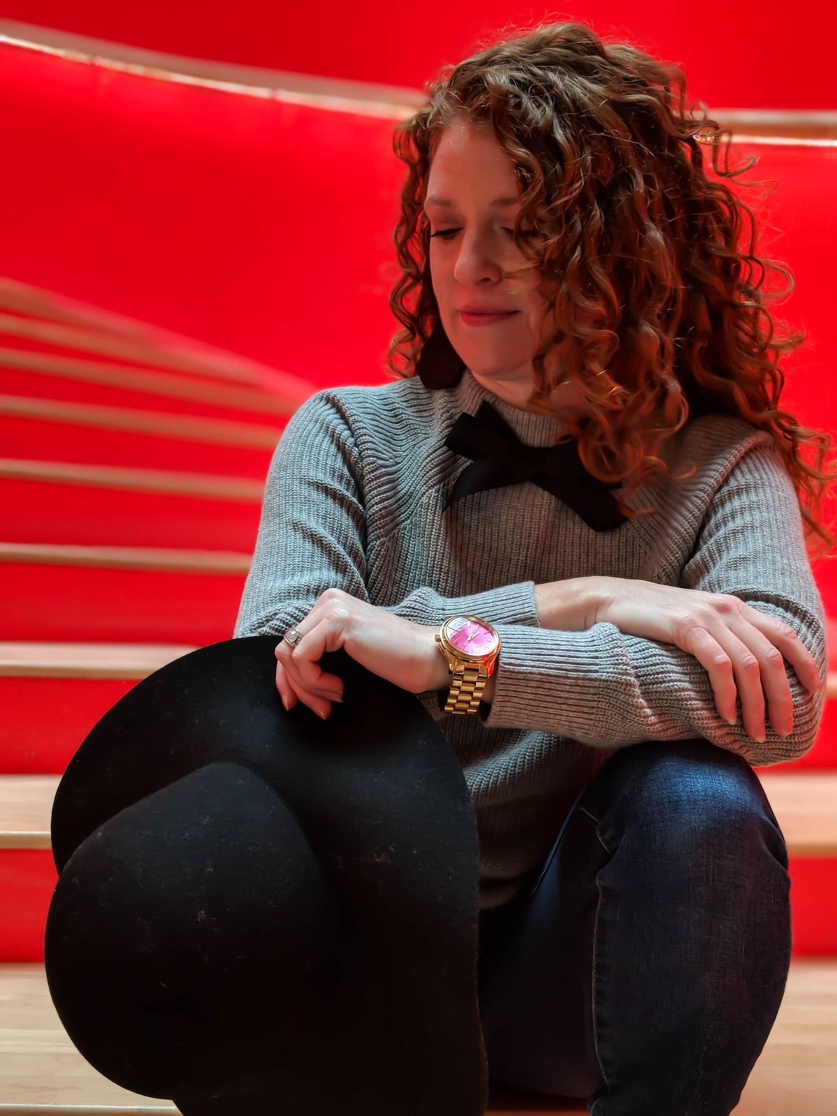 Jamie is sitting on red staircase at the Crosstown Concourse. Close up of me looking down at my hand that is holding my black floppy hat from Forever 21. I am wearing my J Crew Factory bow sweater, AG Jeans, Nickel and Suede black cork earrings and Michael Kors gold watch with the hot pink face.