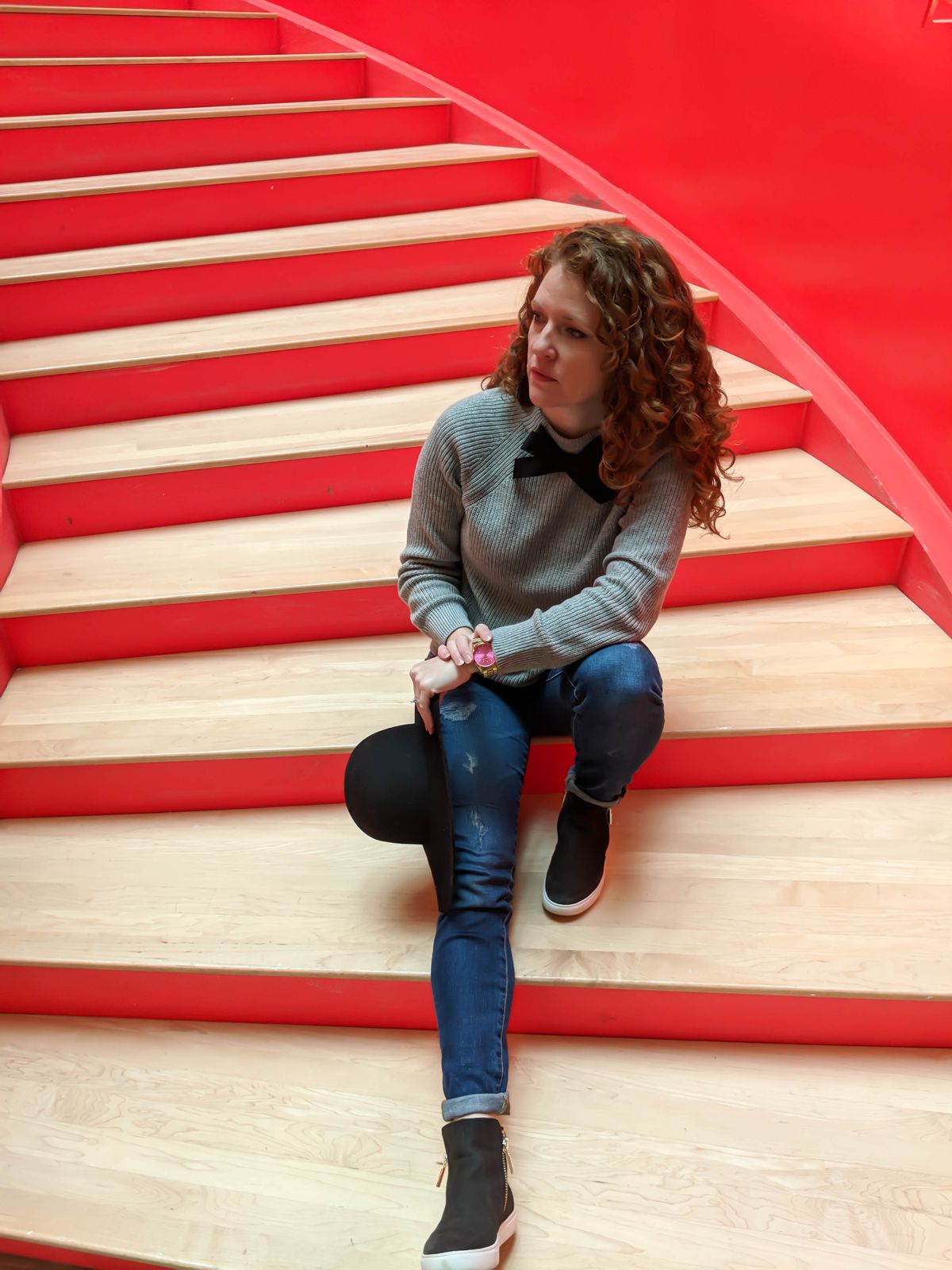 Sitting on the red stair case at Crosstown Concourse with my J Crew Factory Bow Sweater on with AG jeans and sneakers and I'm holding at floppy hat.