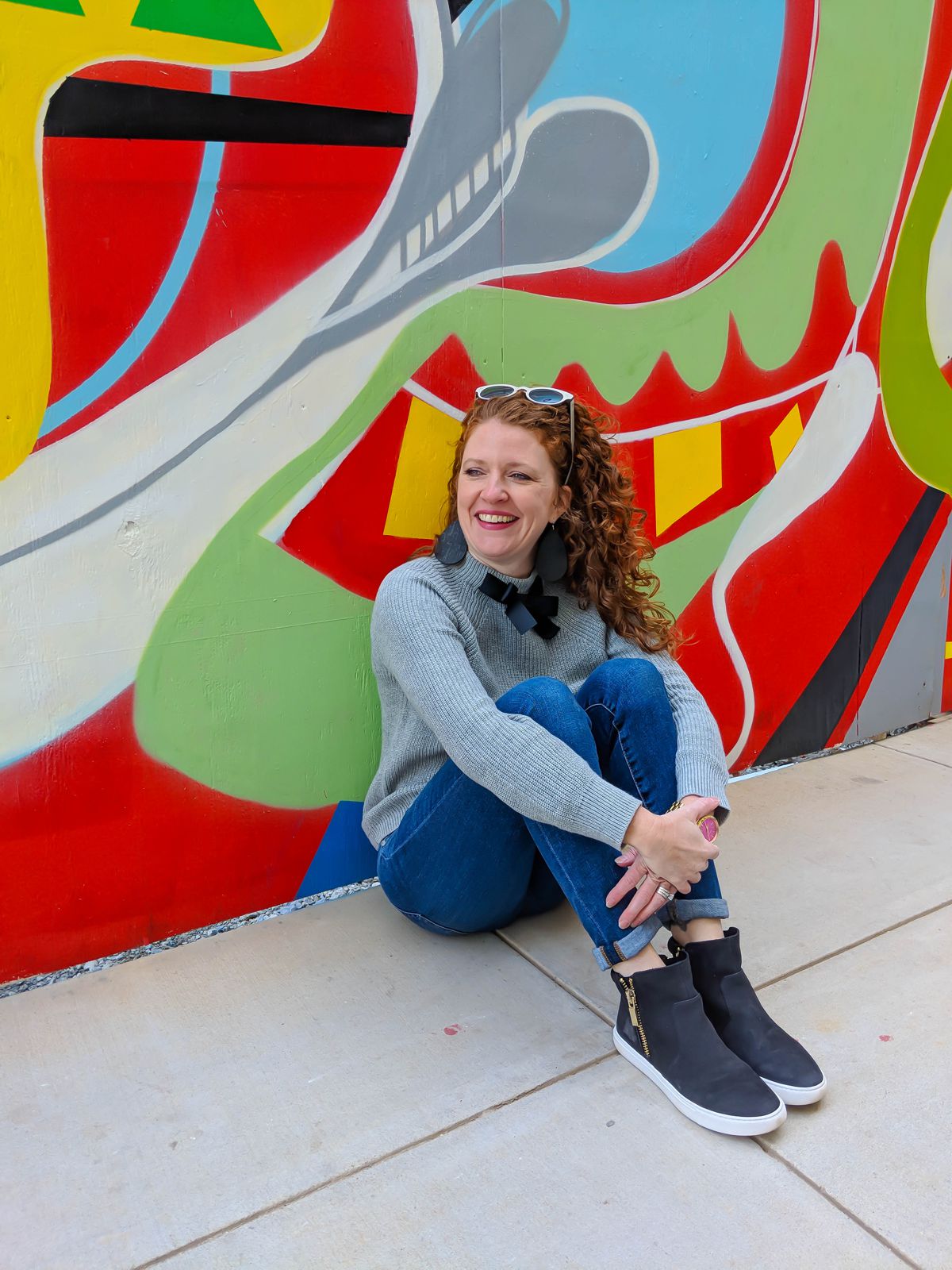 Jamie sitting with knees up and arms around legs at the mural outside of Crosstown Concourse wearing J Crew Factory bow sweater, AG jeans, Kenneth Cole black sneakers, SALT Lorna sunglasses and Nickel and Sued black cork earrings.