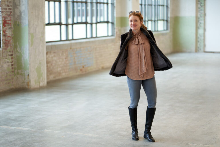 Jamie standing with hands in pockets of faux fur vest paired with a Zara print blouse with a bow and gray jeans and black knee high boots. Wearing chunky stud earrings and Salt Chunky Lorna sunglasses