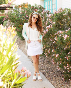 Jamie is standing by flowers looking at camera and crossing her legs. She is wearing spring blazer outfit