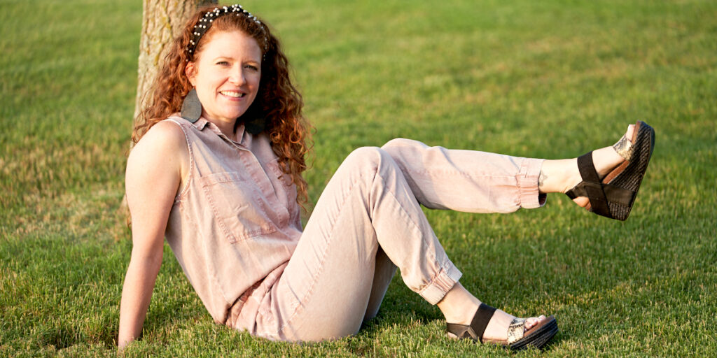 Jamie sitting in the grass with her foot kicked up showing off the shoes. Wearing a pink jumpsuit and OTBT Bushnell Sandals