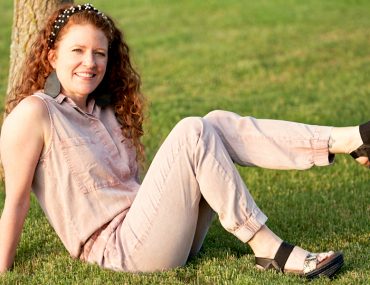Jamie sitting in the grass with her foot kicked up showing off the shoes. Wearing a pink jumpsuit and OTBT Bushnell Sandals