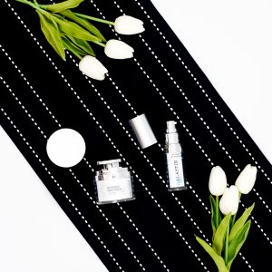 Flat lay of products from Alastin laying on a dark blue and white striped cloth and some white tulips in each side of the products